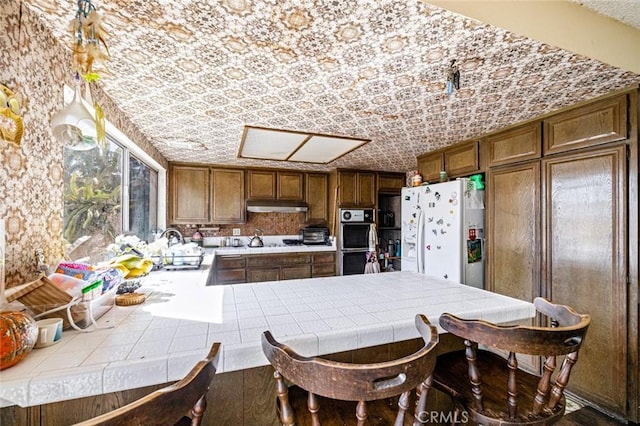 kitchen featuring double oven, under cabinet range hood, a peninsula, tile counters, and white fridge with ice dispenser