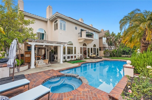 back of house featuring a balcony, a pool with connected hot tub, stucco siding, a chimney, and a patio area