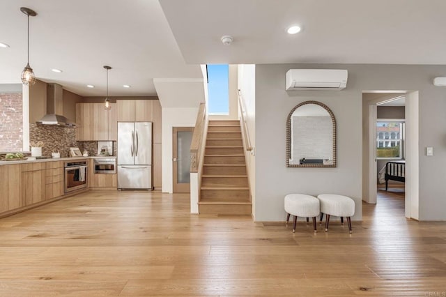 kitchen with a wall unit AC, appliances with stainless steel finishes, light brown cabinets, wall chimney range hood, and modern cabinets