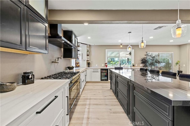 kitchen featuring wall chimney exhaust hood, light wood-style flooring, light stone countertops, stainless steel stove, and white cabinetry