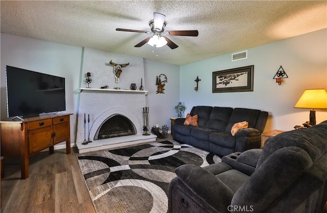 living room with a textured ceiling, a fireplace with raised hearth, dark wood-type flooring, visible vents, and a ceiling fan