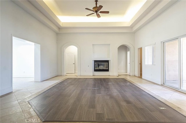 unfurnished living room with ceiling fan, a high ceiling, a tray ceiling, and a glass covered fireplace