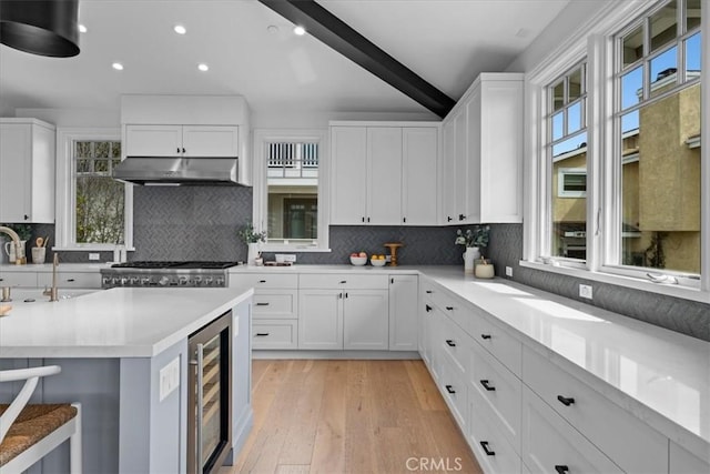 kitchen featuring beam ceiling, light countertops, white cabinetry, beverage cooler, and under cabinet range hood