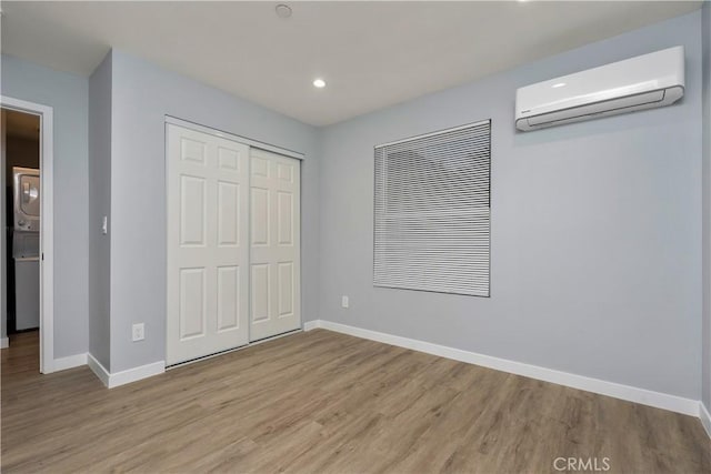 unfurnished bedroom featuring a wall unit AC, stacked washer / drying machine, a closet, light wood-type flooring, and baseboards