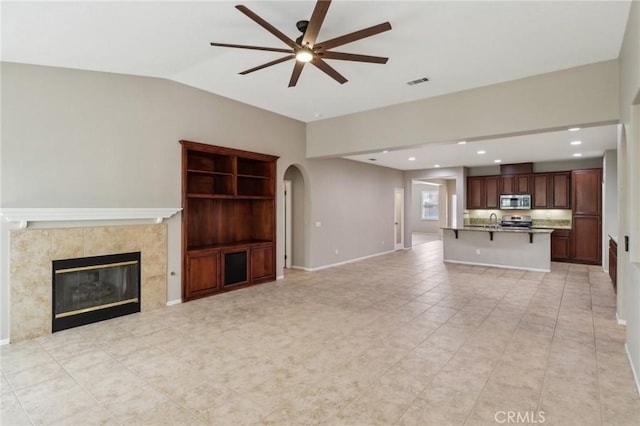 unfurnished living room with ceiling fan, arched walkways, visible vents, vaulted ceiling, and a tiled fireplace