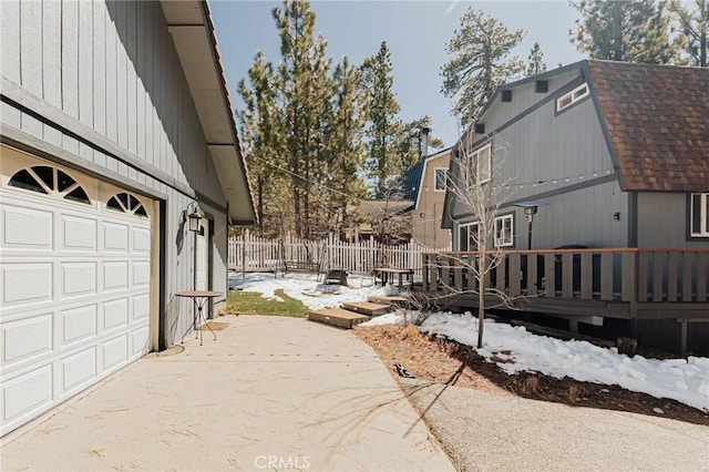 view of snow covered exterior featuring a garage, concrete driveway, roof with shingles, and fence