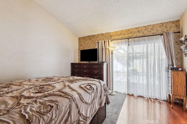 bedroom with wallpapered walls, wood finished floors, access to outside, vaulted ceiling, and a textured ceiling