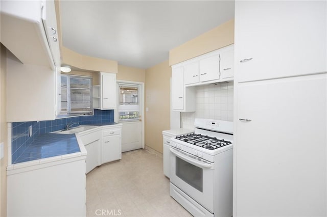 kitchen with tile countertops, white gas stove, a sink, white cabinetry, and backsplash