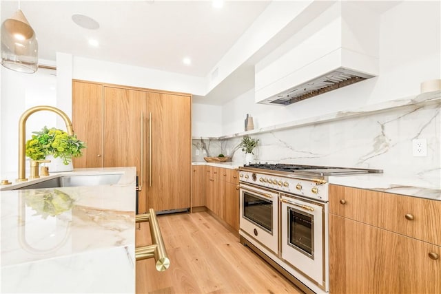 kitchen featuring range with two ovens, light wood finished floors, custom range hood, backsplash, and a sink