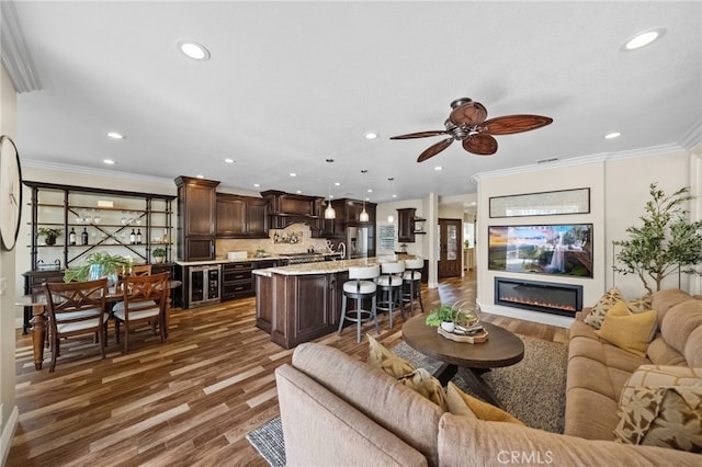 living room featuring dark wood finished floors, a glass covered fireplace, ceiling fan, crown molding, and recessed lighting