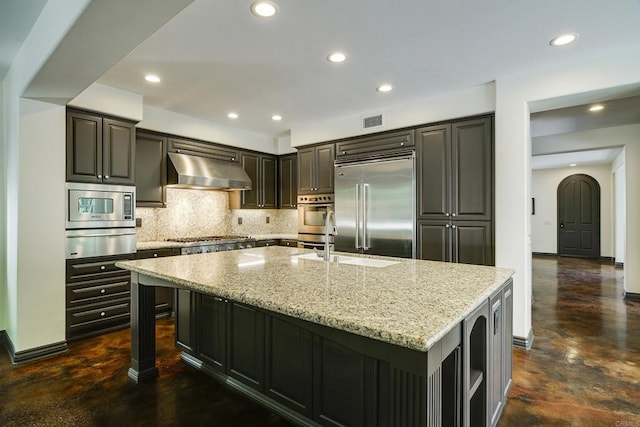 kitchen featuring range hood, light stone countertops, visible vents, a large island with sink, and built in appliances