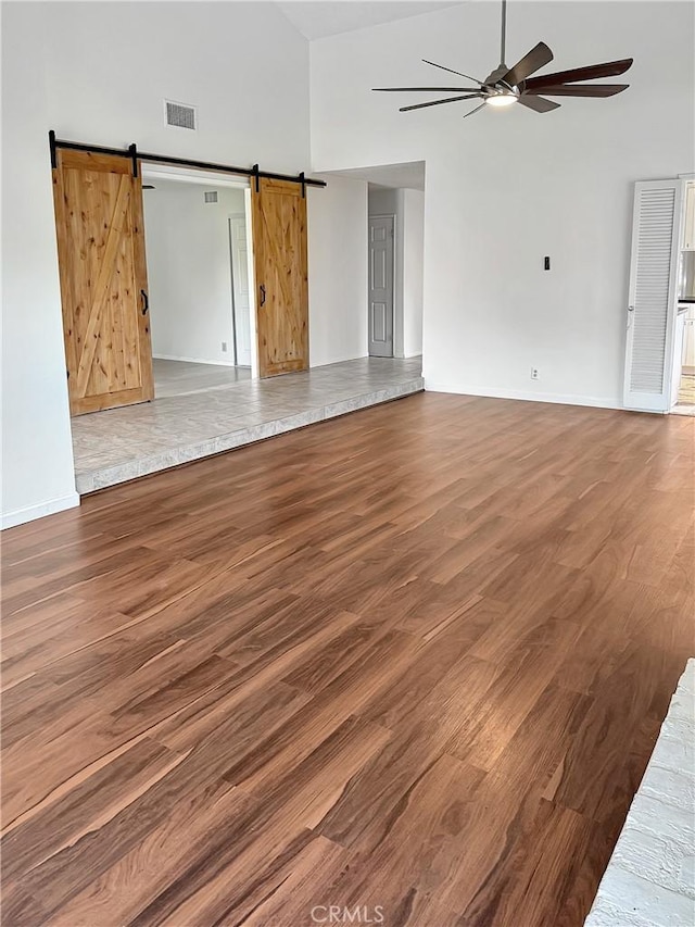 unfurnished living room with visible vents, a barn door, a ceiling fan, wood finished floors, and high vaulted ceiling