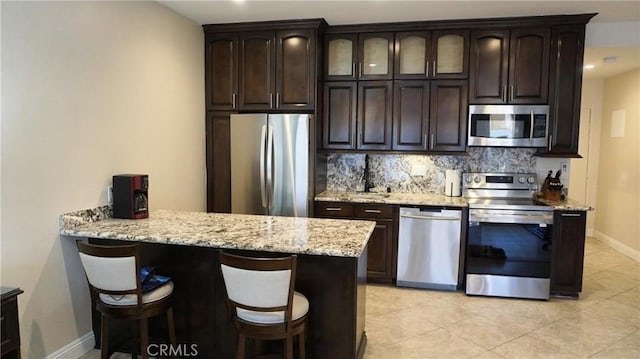 kitchen with light stone countertops, stainless steel appliances, dark brown cabinets, a kitchen bar, and backsplash