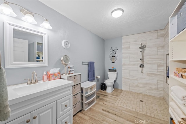 bathroom with a textured ceiling, toilet, wood finished floors, vanity, and a tile shower