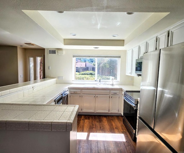 kitchen with tile counters, a raised ceiling, appliances with stainless steel finishes, dark wood-type flooring, and a peninsula