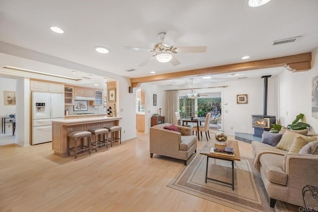 living area featuring beam ceiling, visible vents, light wood-style flooring, a ceiling fan, and a wood stove