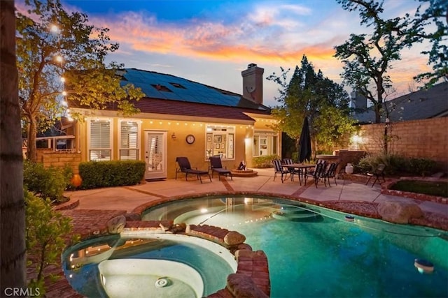 back of house at dusk featuring fence, a pool with connected hot tub, roof mounted solar panels, a chimney, and a patio area