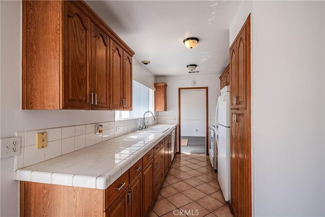 kitchen featuring brown cabinets, tile counters, freestanding refrigerator, light tile patterned flooring, and a sink