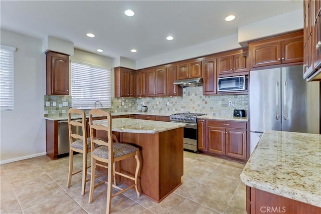 kitchen featuring a breakfast bar, a center island, stainless steel appliances, light stone countertops, and under cabinet range hood