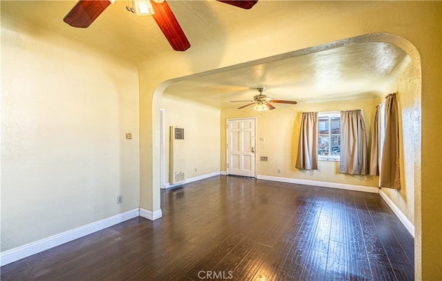 empty room with arched walkways, dark wood-style flooring, visible vents, ceiling fan, and baseboards