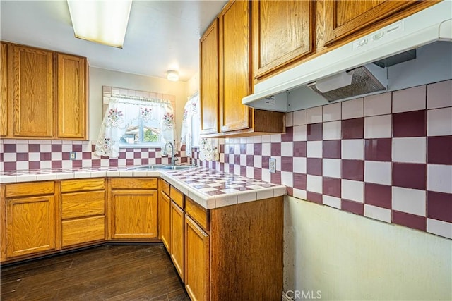 kitchen featuring decorative backsplash, brown cabinets, dark wood-style flooring, under cabinet range hood, and a sink