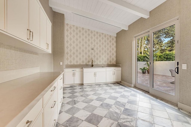 kitchen with white cabinetry, vaulted ceiling with beams, wallpapered walls, and a sink