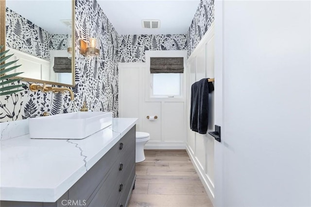 bathroom featuring toilet, wood finished floors, vanity, visible vents, and wallpapered walls