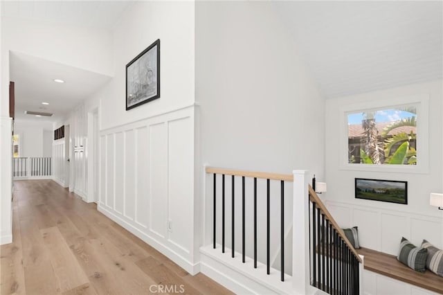 corridor featuring lofted ceiling, a decorative wall, light wood finished floors, and an upstairs landing