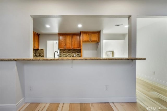 kitchen with decorative backsplash, brown cabinets, a peninsula, light stone countertops, and light wood-style floors