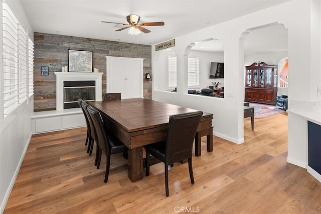 dining room with arched walkways, light wood-style flooring, a large fireplace, ceiling fan, and baseboards