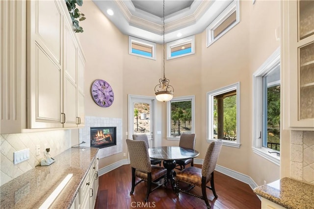 dining space featuring baseboards, a raised ceiling, a tiled fireplace, ornamental molding, and dark wood-type flooring