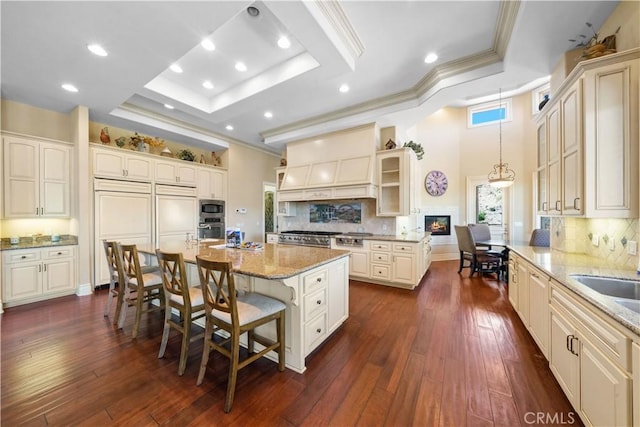 kitchen featuring built in appliances, a kitchen breakfast bar, ornamental molding, a raised ceiling, and custom range hood