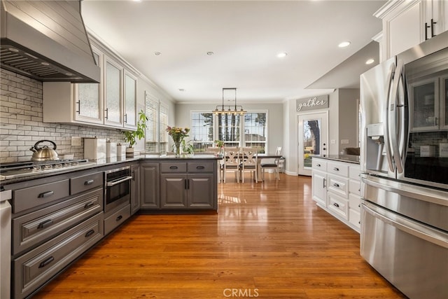 kitchen with appliances with stainless steel finishes, wood finished floors, a peninsula, wall chimney range hood, and backsplash