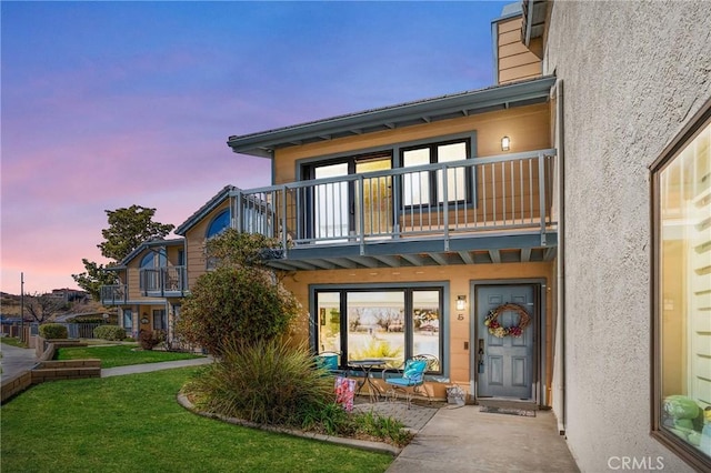 back of property at dusk featuring a yard, a chimney, a balcony, and stucco siding