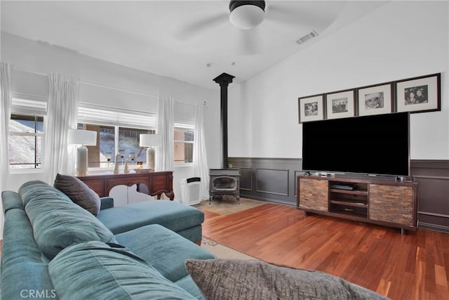 living room featuring a ceiling fan, a wainscoted wall, wood finished floors, and a wood stove