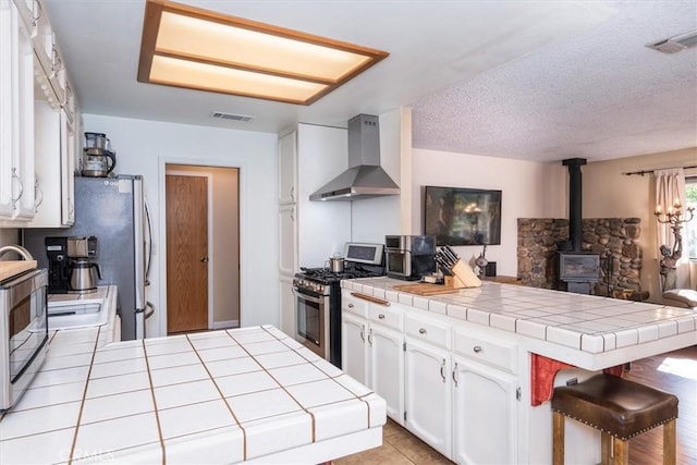 kitchen with a wood stove, wall chimney range hood, a peninsula, and appliances with stainless steel finishes