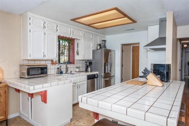 kitchen with white cabinets, appliances with stainless steel finishes, wall chimney range hood, tile counters, and tasteful backsplash