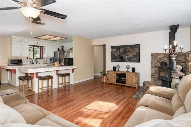 living room with light wood finished floors, visible vents, a ceiling fan, a wood stove, and a textured ceiling