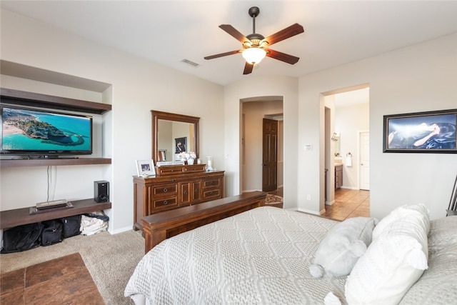 carpeted bedroom with arched walkways, visible vents, ceiling fan, ensuite bath, and baseboards