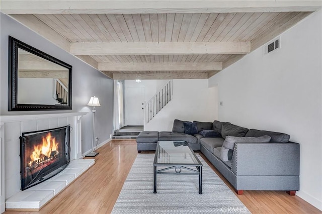 living area featuring a warm lit fireplace, stairs, light wood-style floors, and beamed ceiling