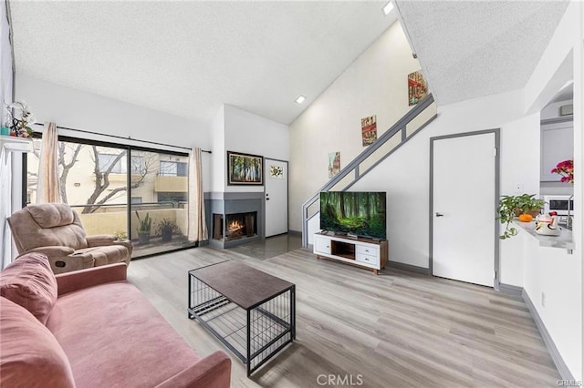 living area with a textured ceiling, high vaulted ceiling, baseboards, a lit fireplace, and light wood-type flooring