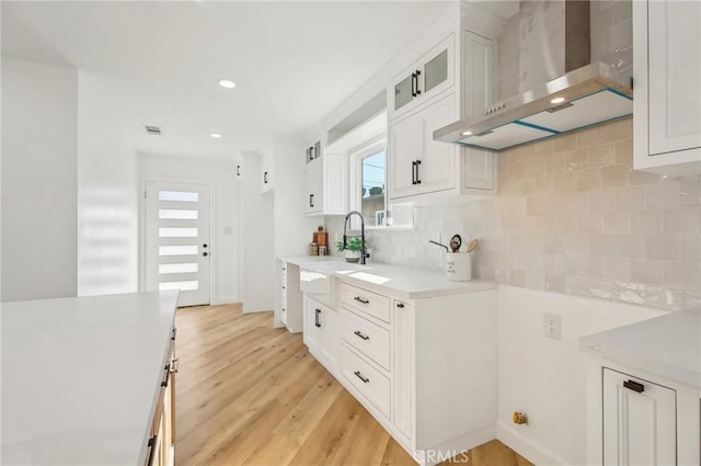 kitchen featuring a sink, light countertops, wall chimney range hood, light wood-type flooring, and tasteful backsplash