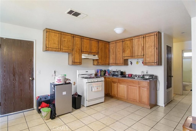 kitchen with light tile patterned flooring, under cabinet range hood, visible vents, brown cabinetry, and white gas range