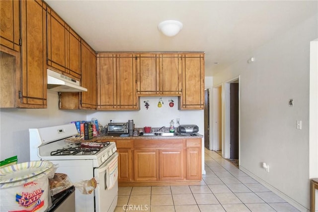 kitchen with light tile patterned floors, white gas range oven, brown cabinetry, under cabinet range hood, and a sink