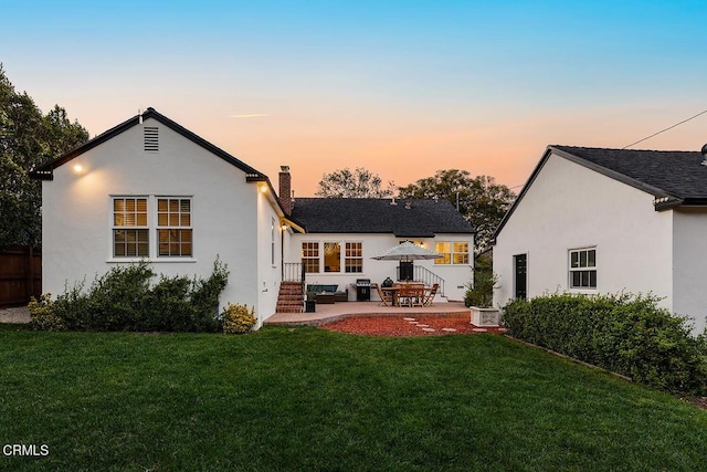 back of house with a shingled roof, a patio, a chimney, a yard, and stucco siding