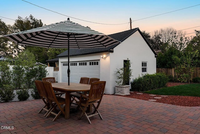 patio terrace at dusk featuring outdoor dining area, an attached garage, and fence