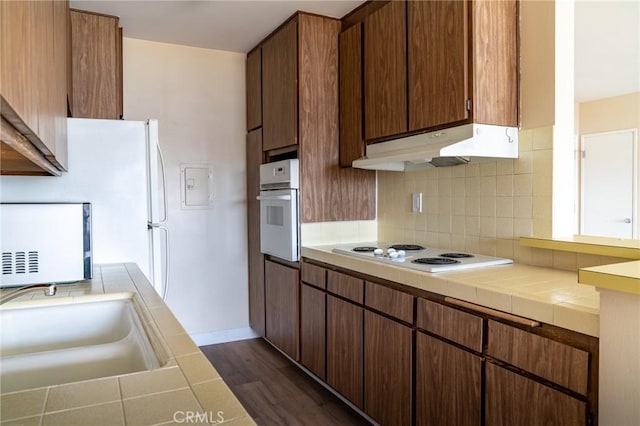 kitchen with white appliances, tile counters, dark wood finished floors, under cabinet range hood, and backsplash