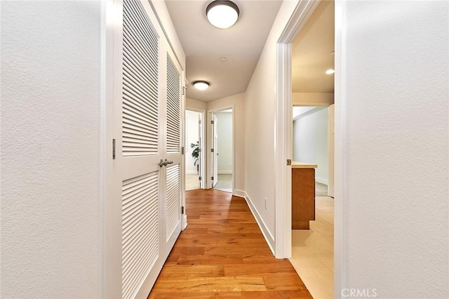 hall with a textured wall, light wood-style flooring, and baseboards