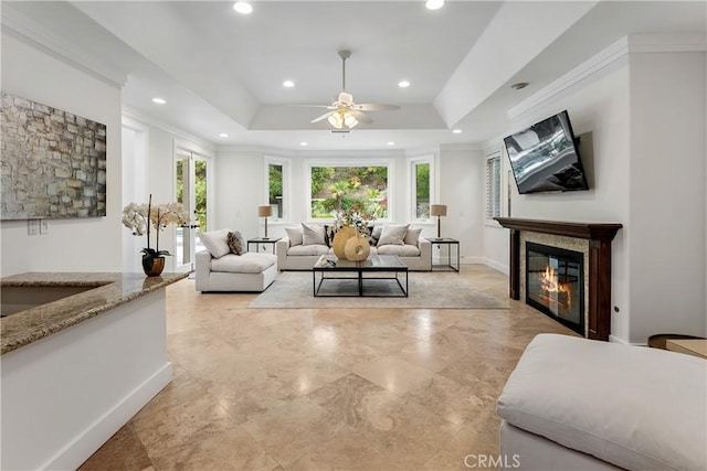 living room with recessed lighting, a tray ceiling, a ceiling fan, crown molding, and a glass covered fireplace