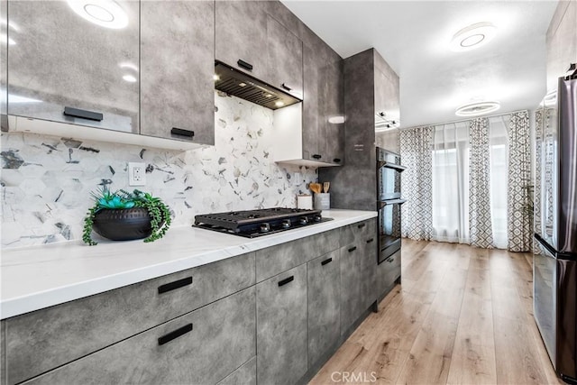 kitchen featuring light countertops, light wood-style flooring, decorative backsplash, ventilation hood, and black appliances